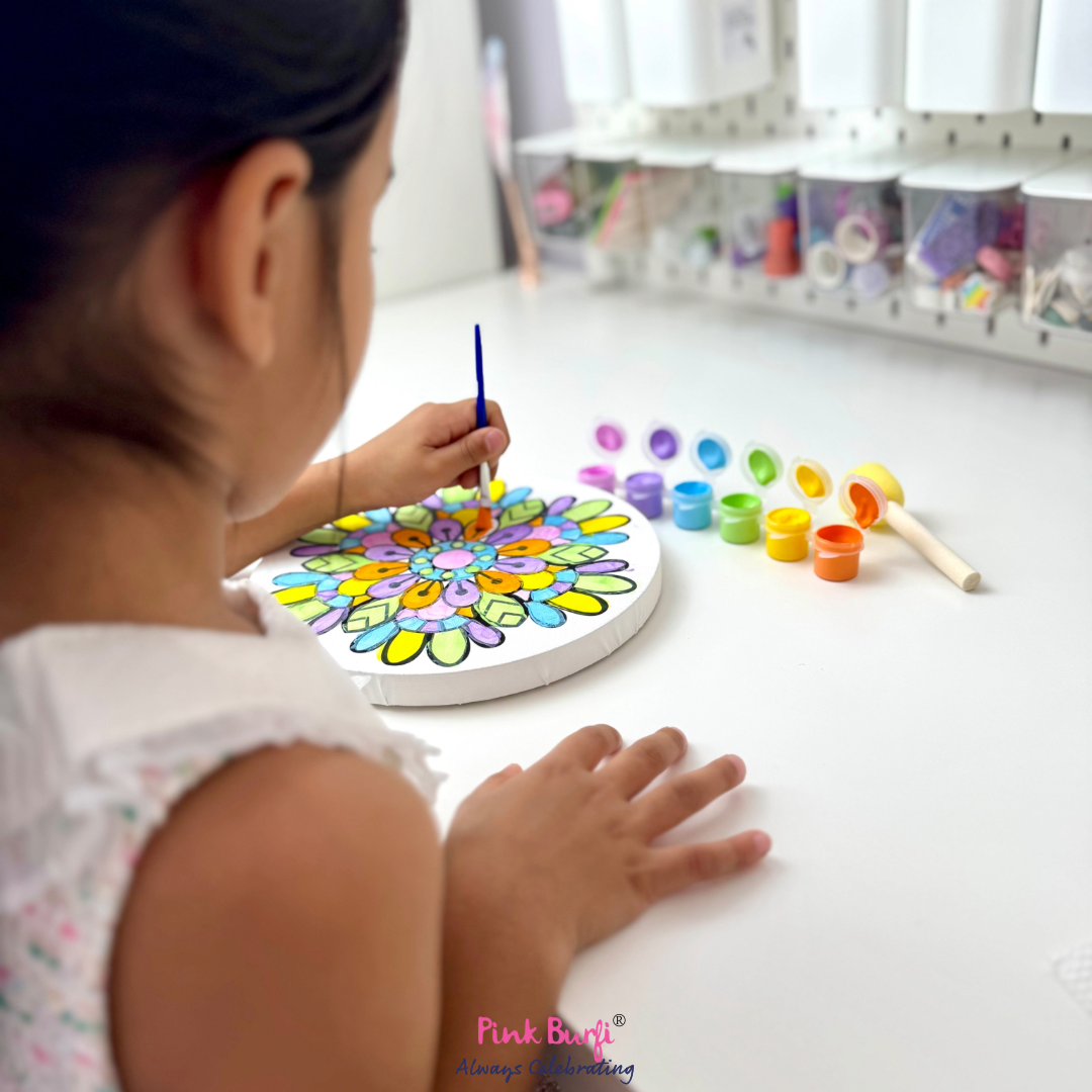 Child painting a colorful mandala on a white board with various colors and tools around from the Pink Burfi Resist Art Canvas Rangoli Box.